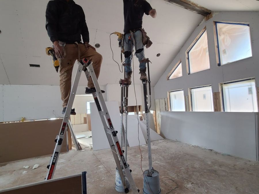 Two L.O.Z. Construction crew members hanging drywall on stilts under a vaulted ceiling
