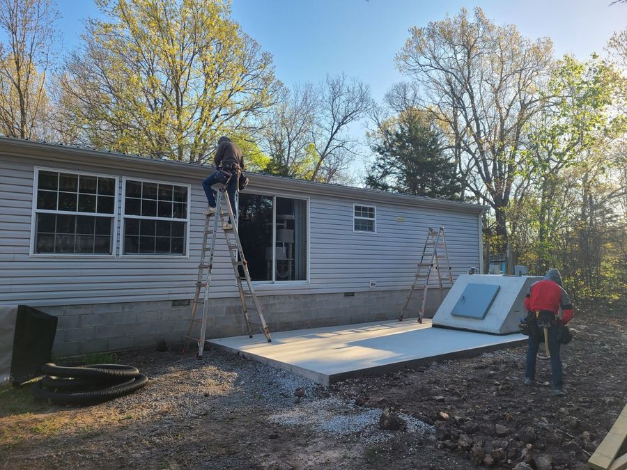 Concrete slab poured with a storm shelter set in place and crew on ladders at the house