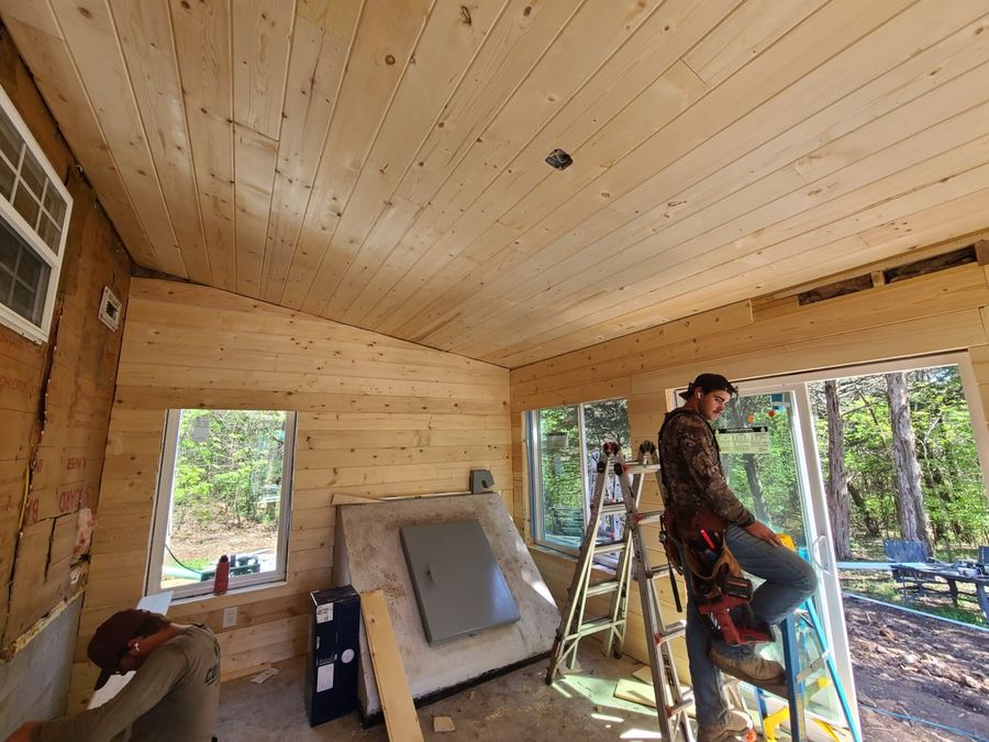 Pine-lined sunroom interior with the storm shelter door and a crew member on a ladder