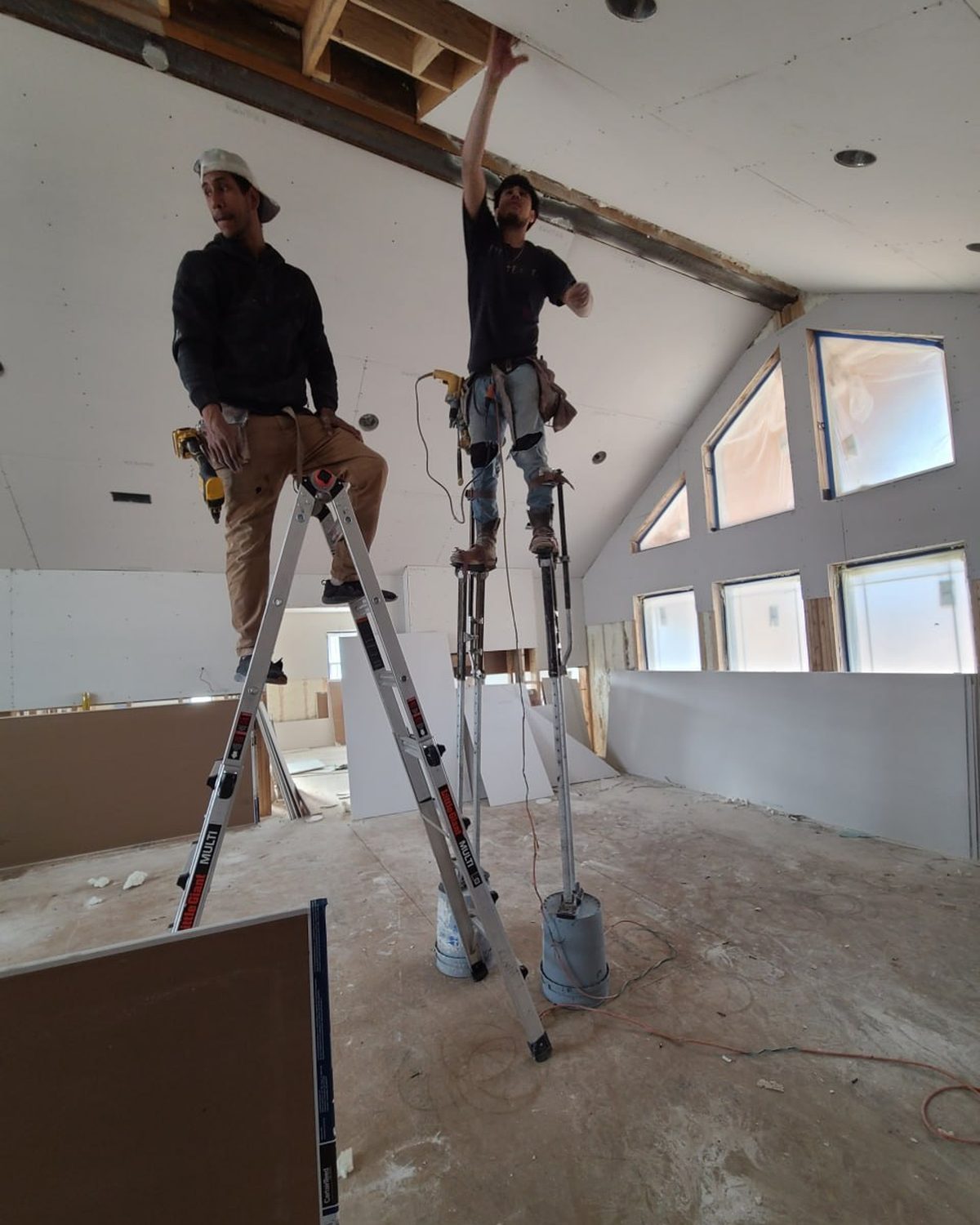 Two L.O.Z. Construction crew members hanging drywall on stilts under a vaulted ceiling